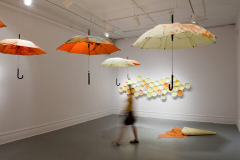 A gallery piece. Umbrellas suspended from the ceiling, a spilled cone with orange liquid on the floor, and round swatches of colour on the wall. Blurred image of woman walking through it.