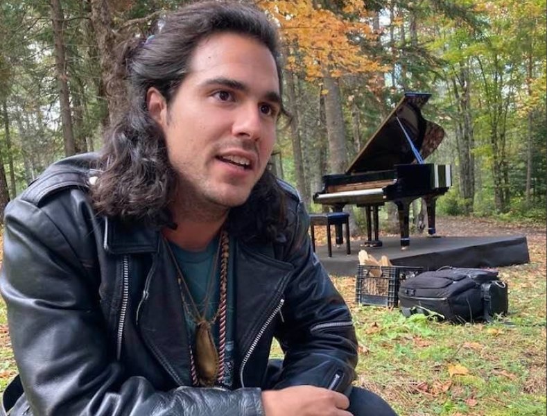Jeremy Dutcher in front of a piano in the forest.