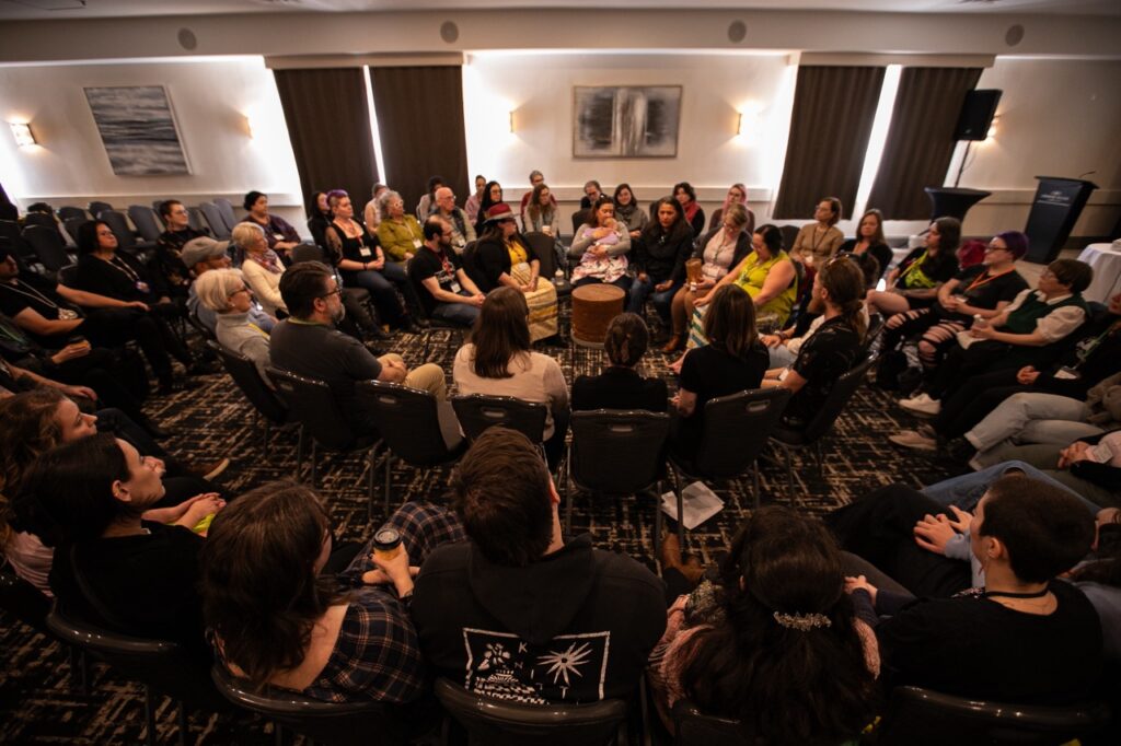 About 50 participants are seated in chairs that are formed into a spiral shape. The artists are seated in the middle, where there is a Wabanaki birch bark basket that is over 100 years old.